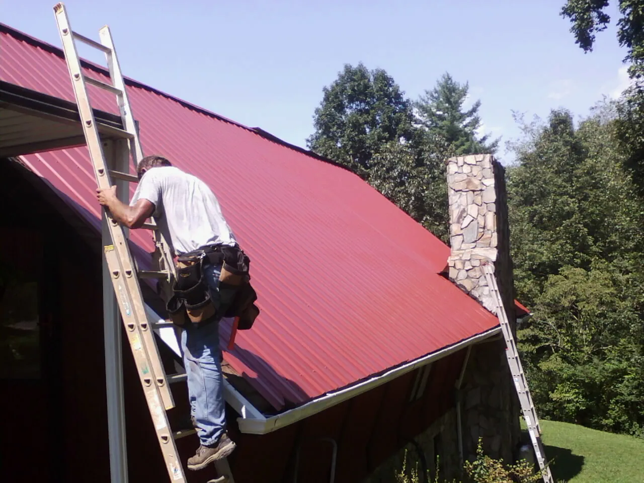 Roofer on a ladder beside a red metal roof and stone chimney — Soffit & Fascia Repair by Roofing Brothers in Blairstown
